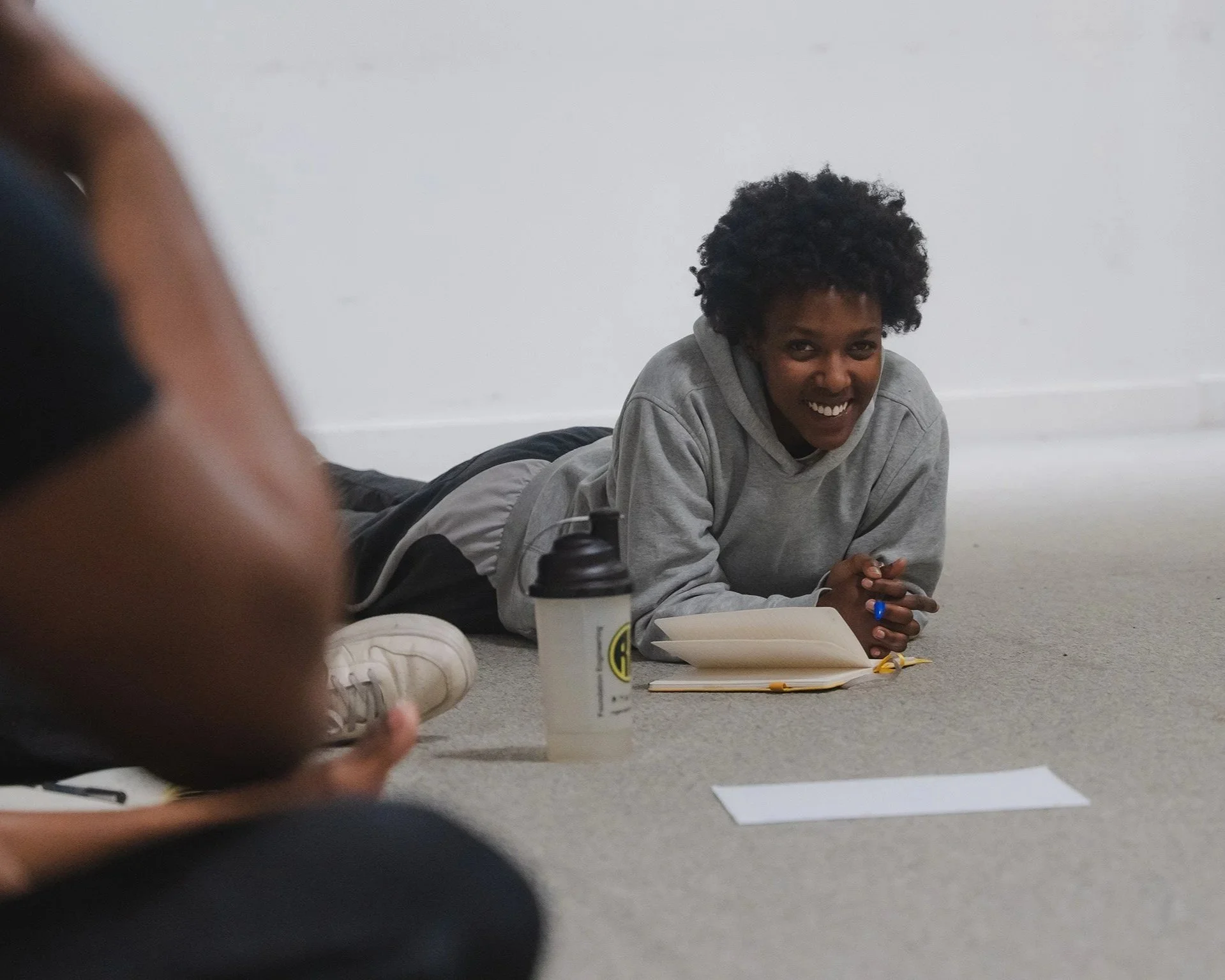 Person lying on the floor smiling, wearing a gray hoodie, with a notebook, a pen, a water bottle, and a piece of paper nearby.