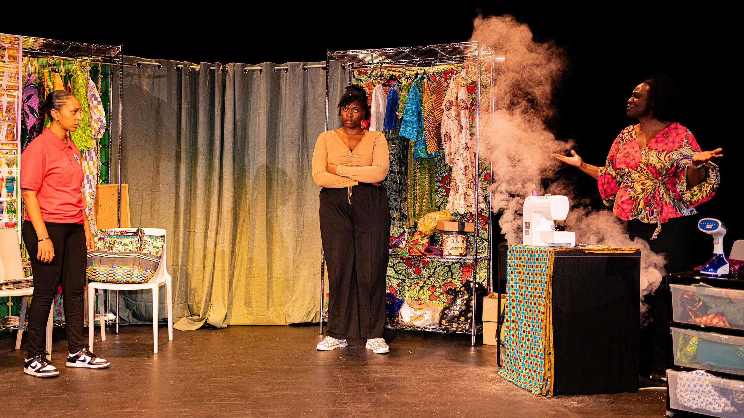 Three women are on stage involved in a theatrical scene; one woman on the left stands in front of a colorful backdrop, another in the middle looks concerned with her arms crossed, and the third on the right is explaining with a cloud of smoke or vapo