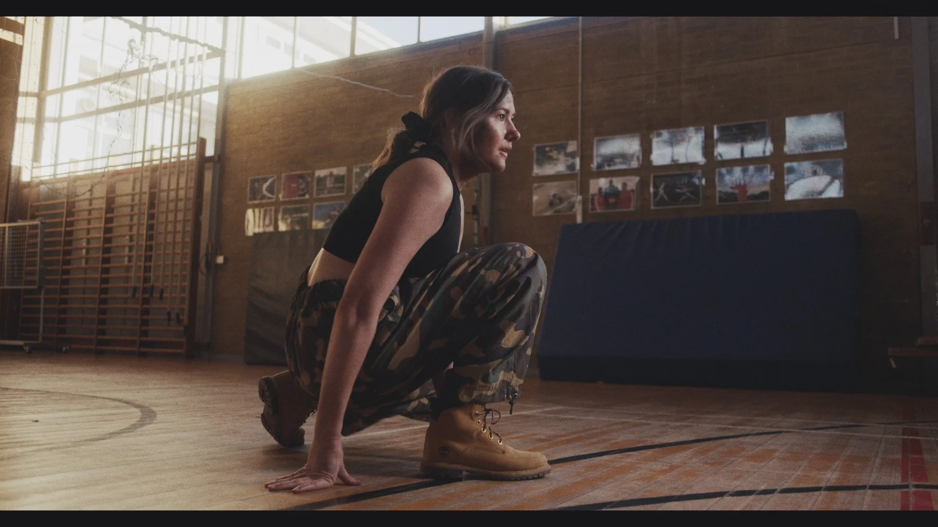 A woman with camouflage pants and tan work boots crouching on an indoor basketball court, with a gymnasium wall and photos in the background.
