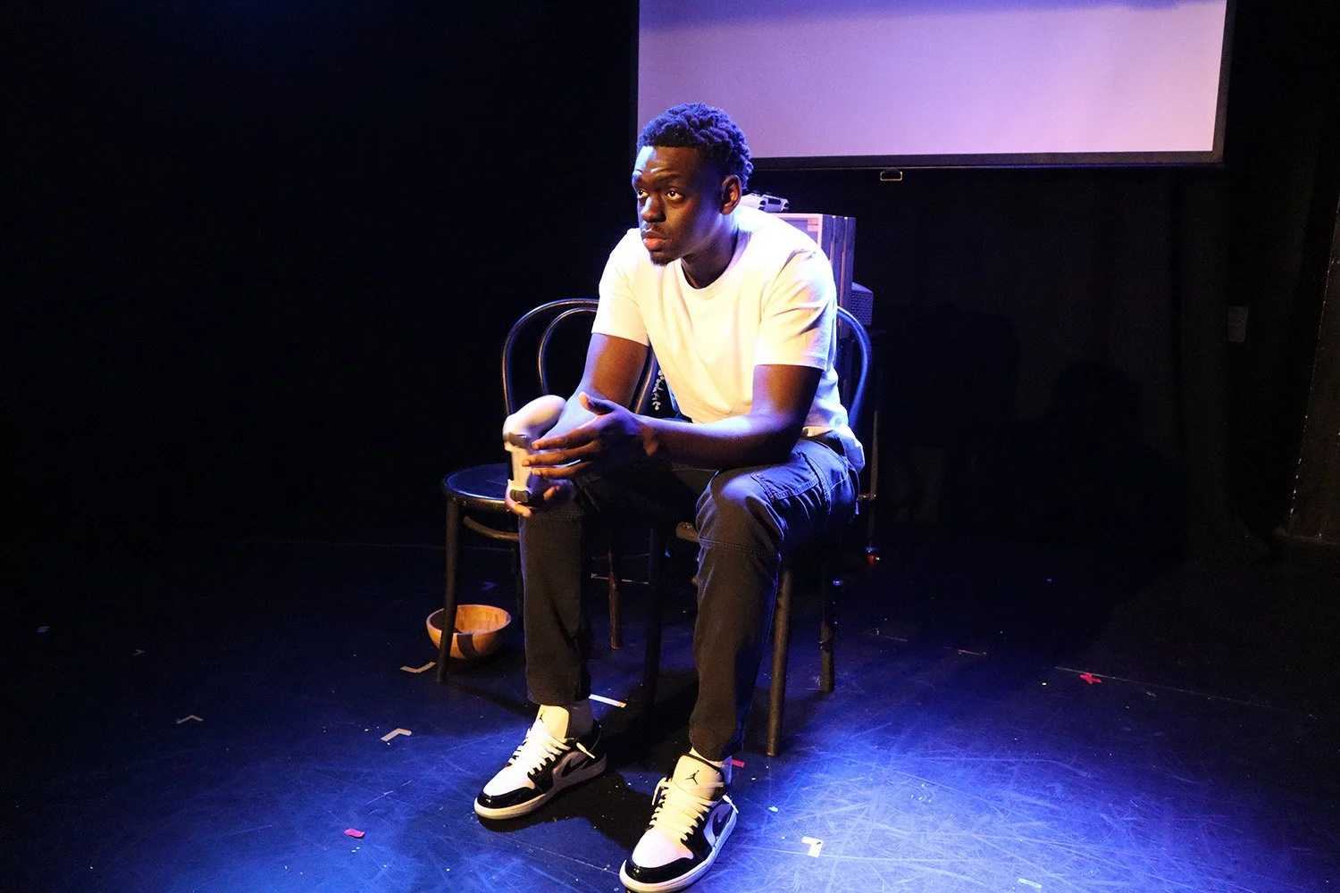 A young man in a white t-shirt, black pants, and black-and-white sneakers sitting on a black chair on a stage with a dark background and a large screen behind him. He is performing in a theatrical production.