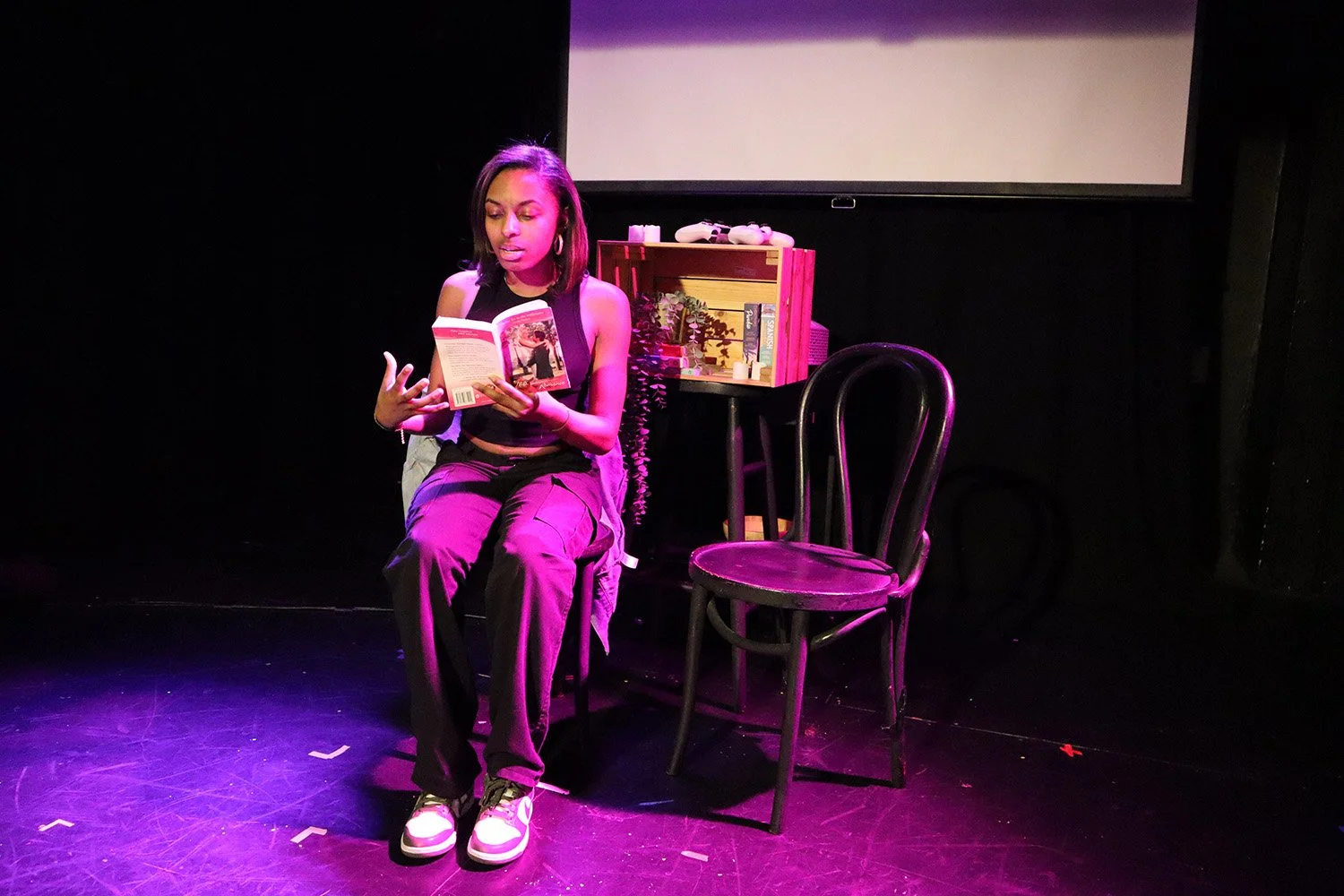 A young woman with shoulder-length black hair, wearing a black sleeveless top and purple pants, is sitting on a black chair on a stage, reading a book. There is a small wooden bookshelf and an empty black chair beside her, with a dark background and 