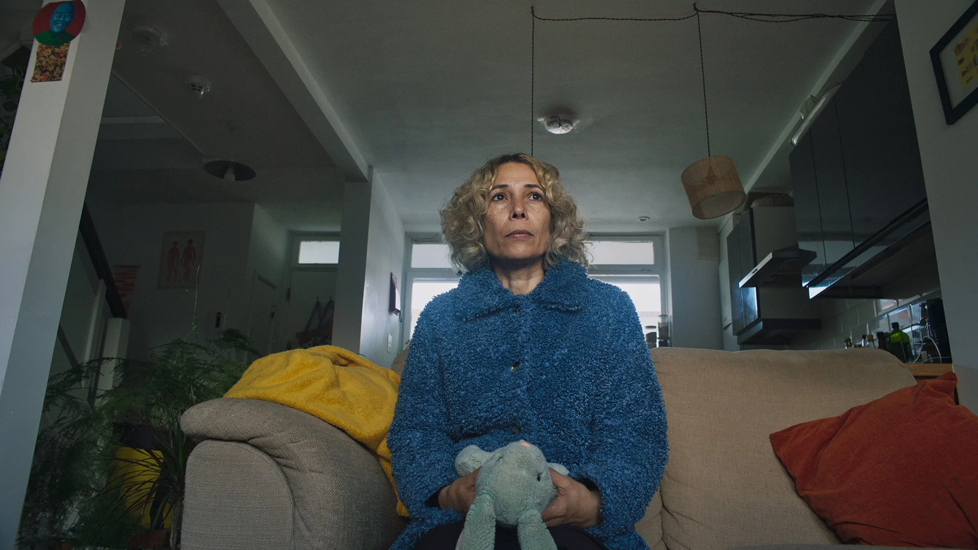 A middle-aged woman with curly blonde hair sitting on a beige couch holding a gray stuffed animal, wearing a blue textured sweater. The living room has a large window behind her, with kitchen cabinets visible to the right, and a yellow blanket on the