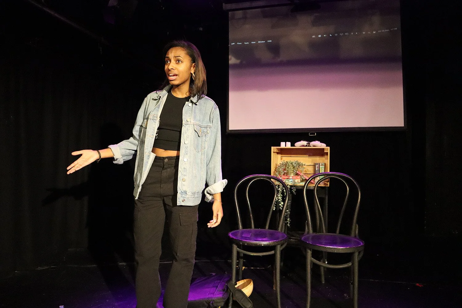 A woman on stage speaking with two black chairs behind her, a small table with plants and books, a large projector screen, all set in a black background stage.