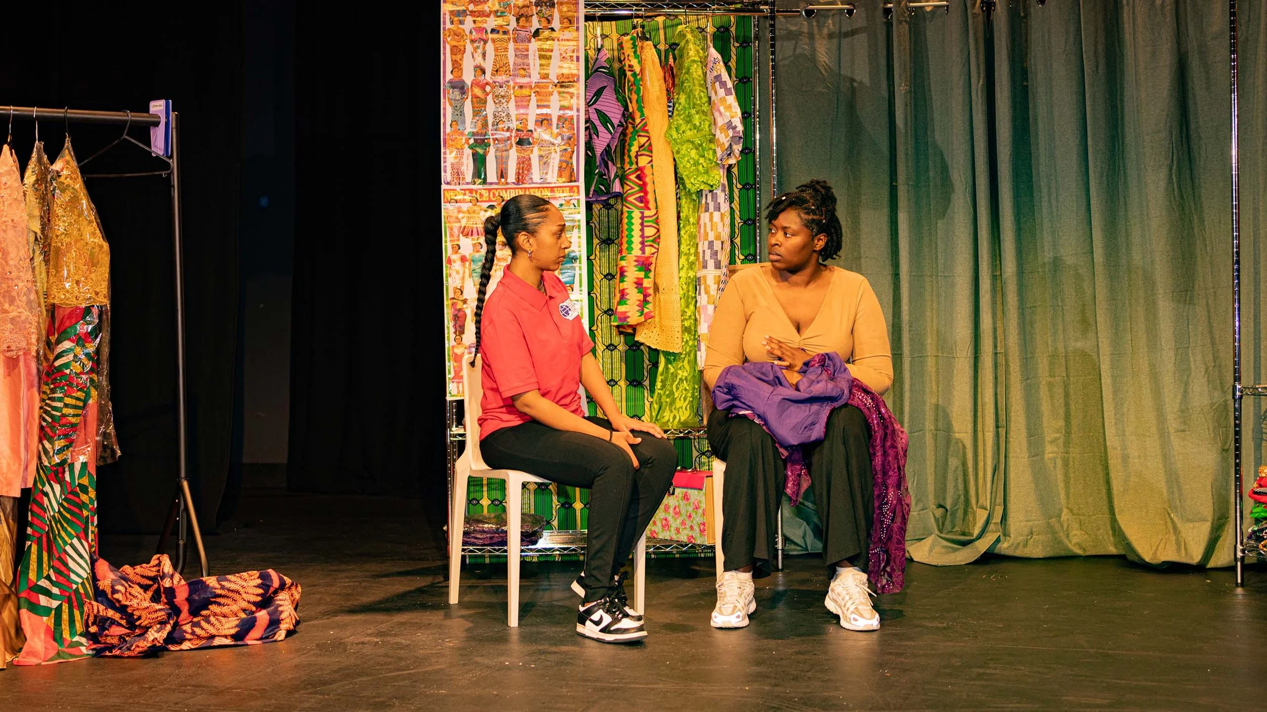 Two women sitting and talking on stage, surrounded by colorful clothing and fabric displays, with a black curtain and green backdrop.