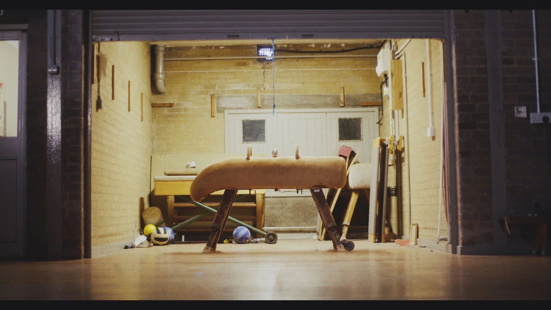 A garage with various sports equipment and wooden furniture, illuminated by indoor lighting, with brick walls and a concrete floor.