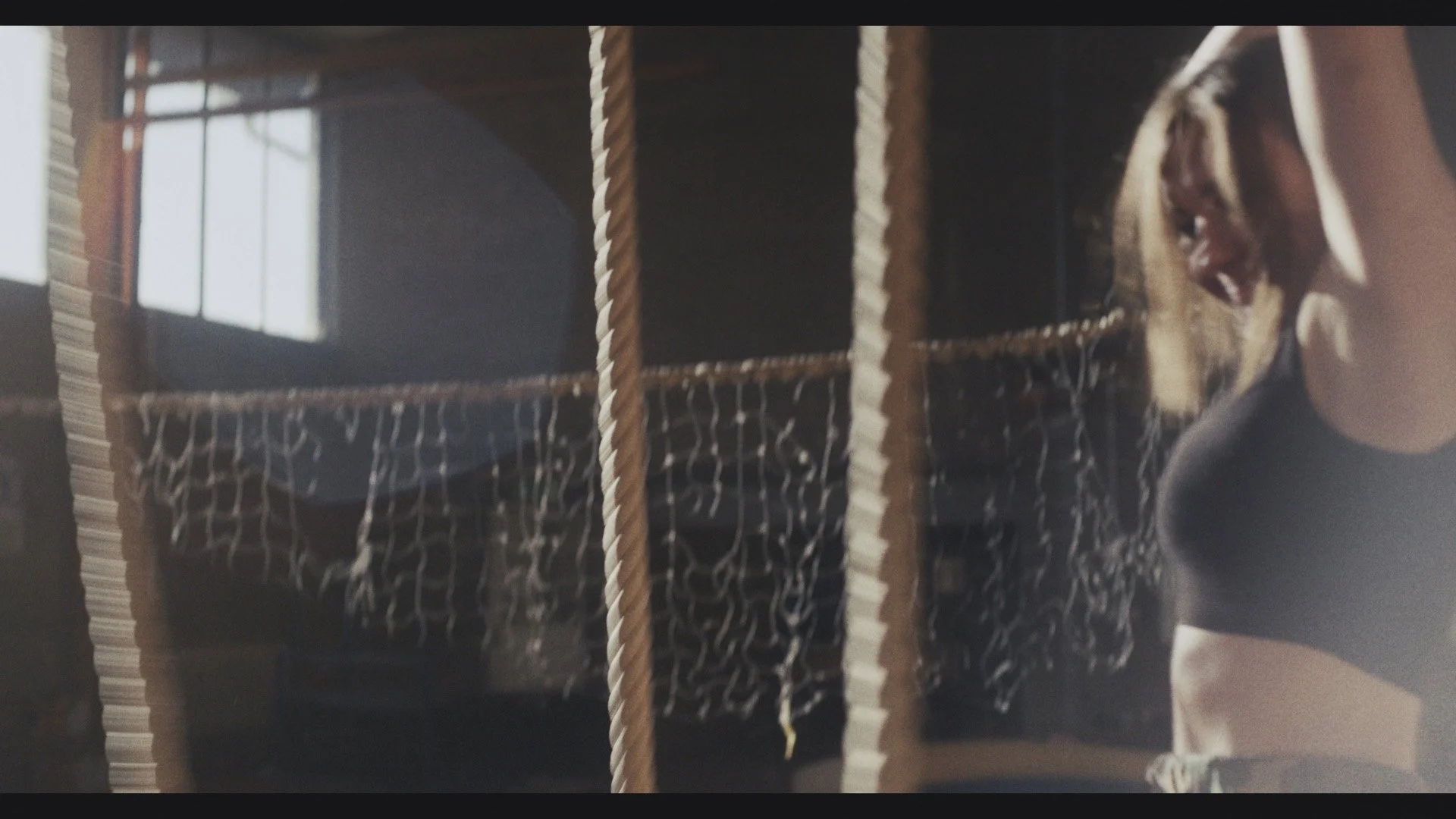 A woman with blonde hair in a ponytail wearing a black t-shirt, avoids gym ropes swinging in front of her.