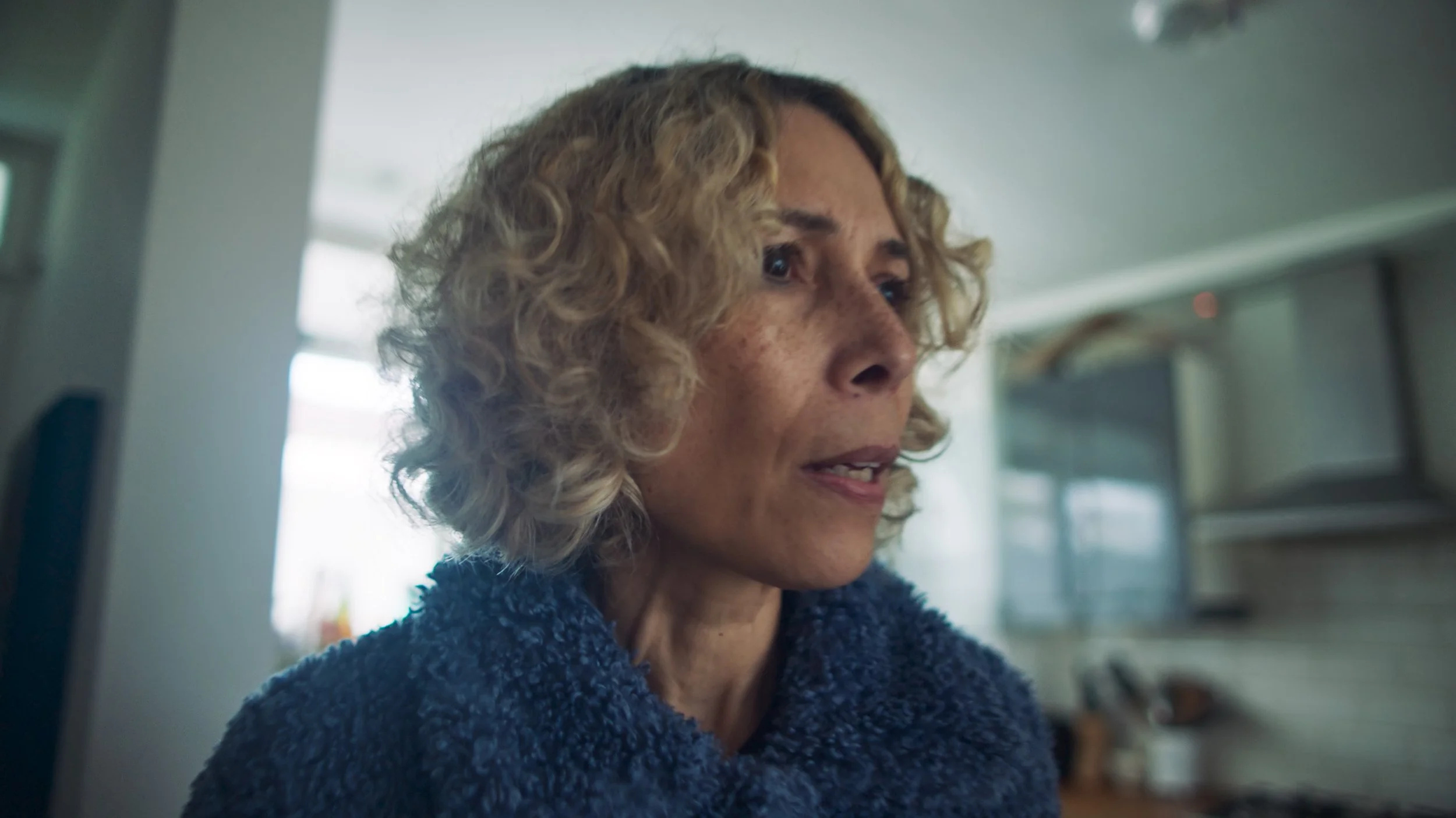 Close-up of a woman with curly blonde hair and freckles, wearing a blue, fuzzy sweater, indoors with a kitchen background.