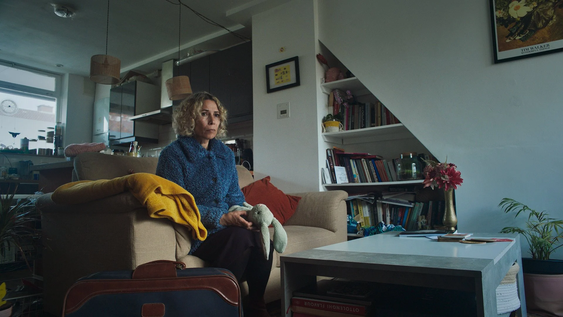 A woman with curly blonde hair sitting on a beige couch in a living room, holding a stuffed rabbit, with a yellow blanket and red pillow beside her. There are bookshelves, plants, and wall art in the background.