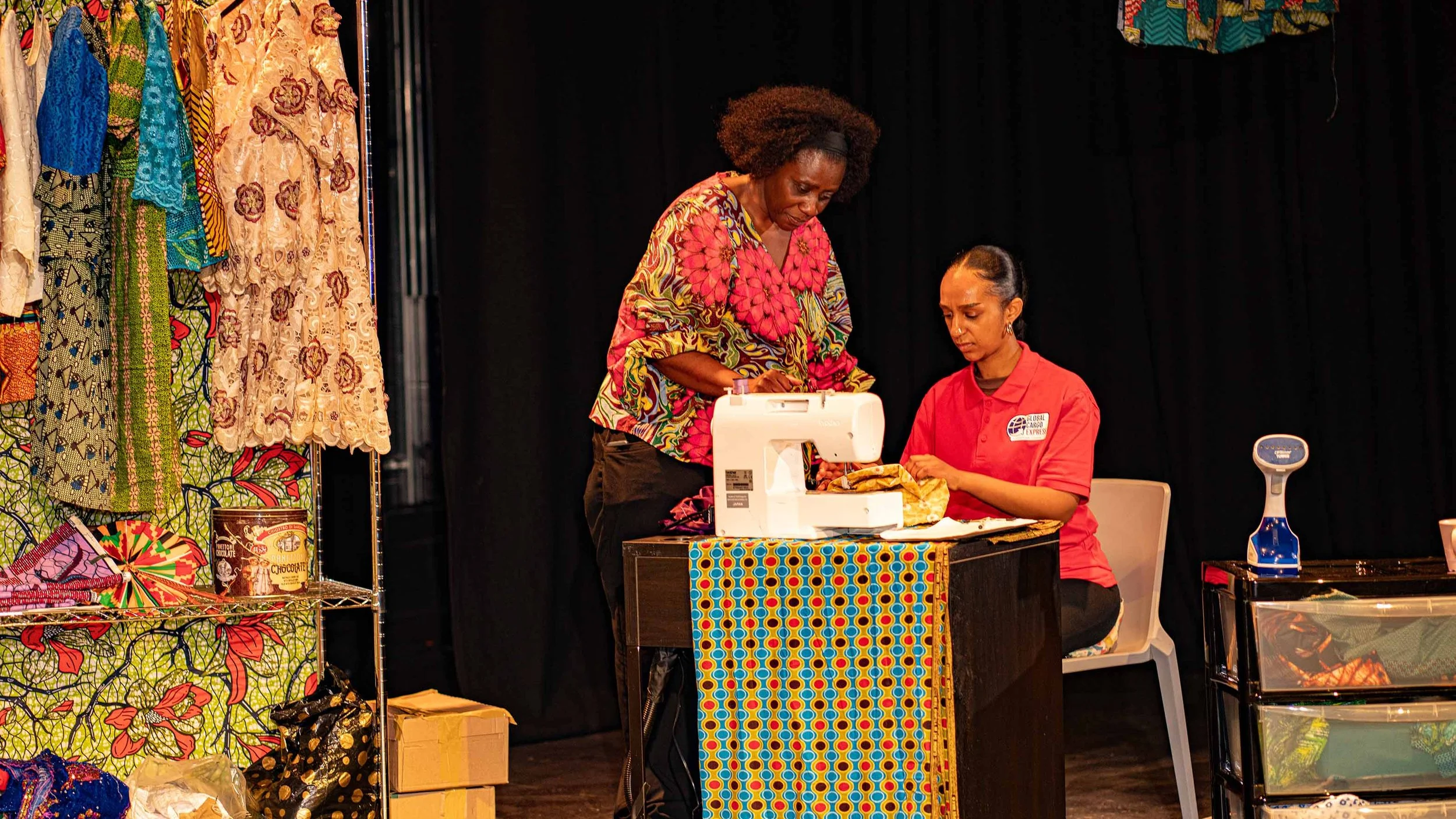 Two women are on stage involved in a theatrical scene; they are sewing with colourful fabric and clothing around them on stage.