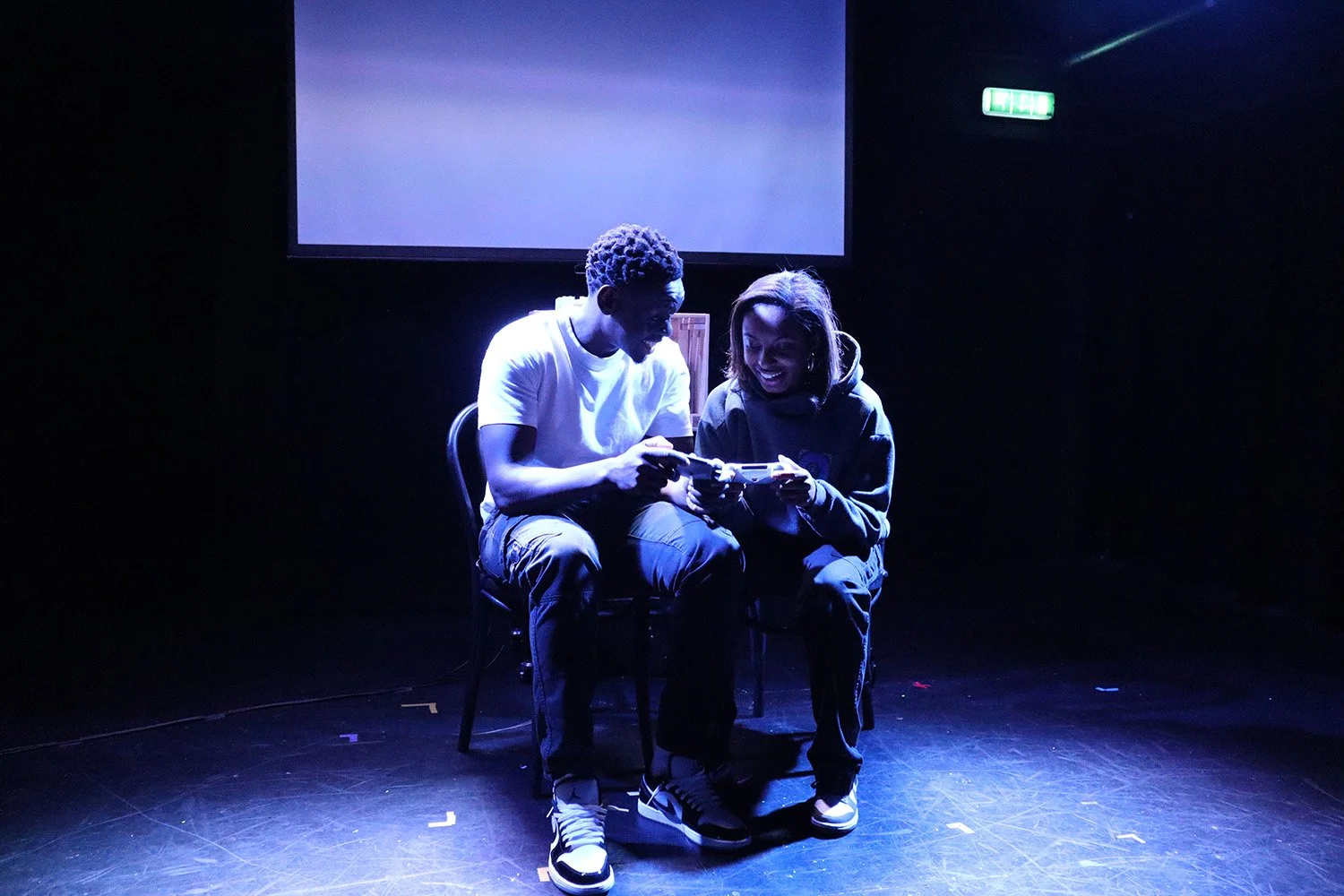 Two people sitting on stage chairs, as part of a theatrical performance, playing video games together, with smiles, illuminated by blue stage lights, with a large blank screen behind them, in a dark room.
