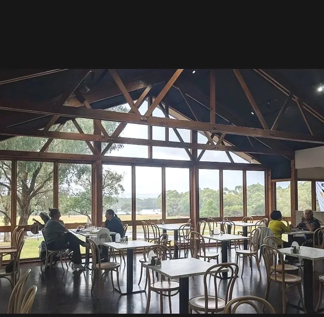 A spacious restaurant with large glass windows overlooking trees and water. Wooden chairs and white tables are arranged throughout, with some patrons seated and conversing.