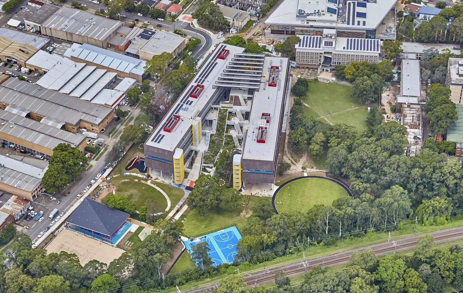 Aerial view of a modern multi-story building with outdoor sports courts, green fields, and surrounding trees.