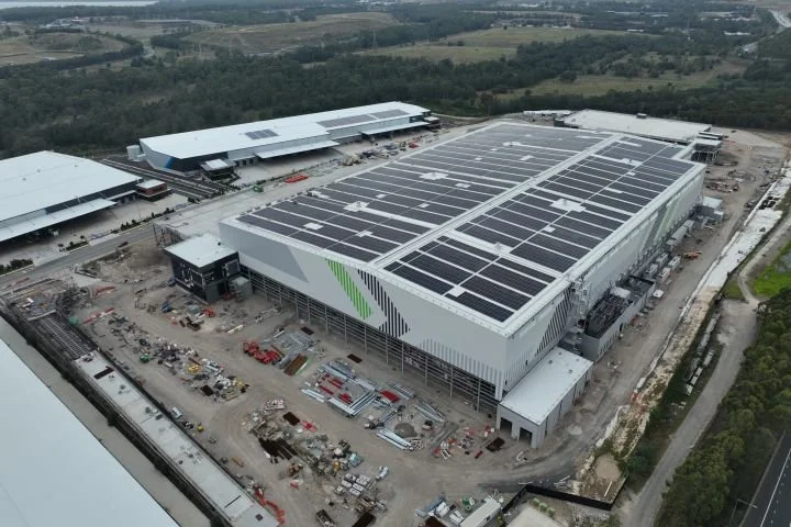 Aerial view of a large commercial building under construction with solar panels on the roof. Nearby, there are other industrial buildings and a developing area with construction equipment and materials.