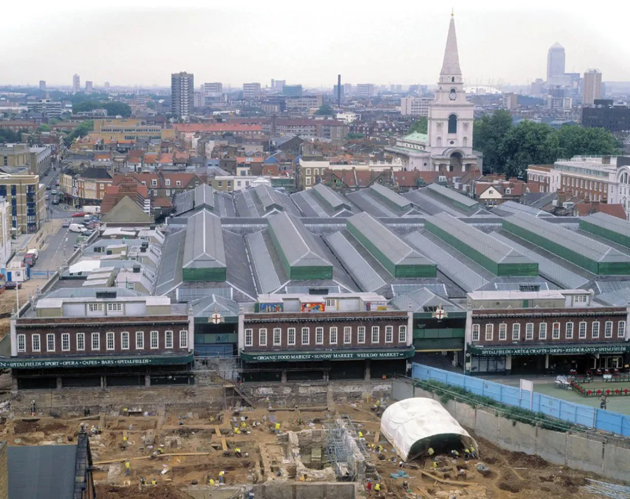 Aerial cityscape view showing a historic market building with a large roof, a church with a tall steeple, and ongoing construction work in the foreground.
