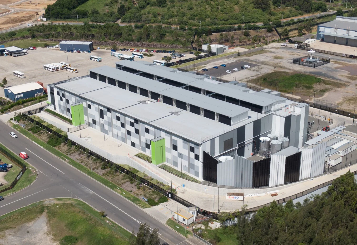 A large, modern building with a gray and black checkered exterior, green doors, and a flat roof under construction, surrounded by paved roads, parking lots, and greenery.