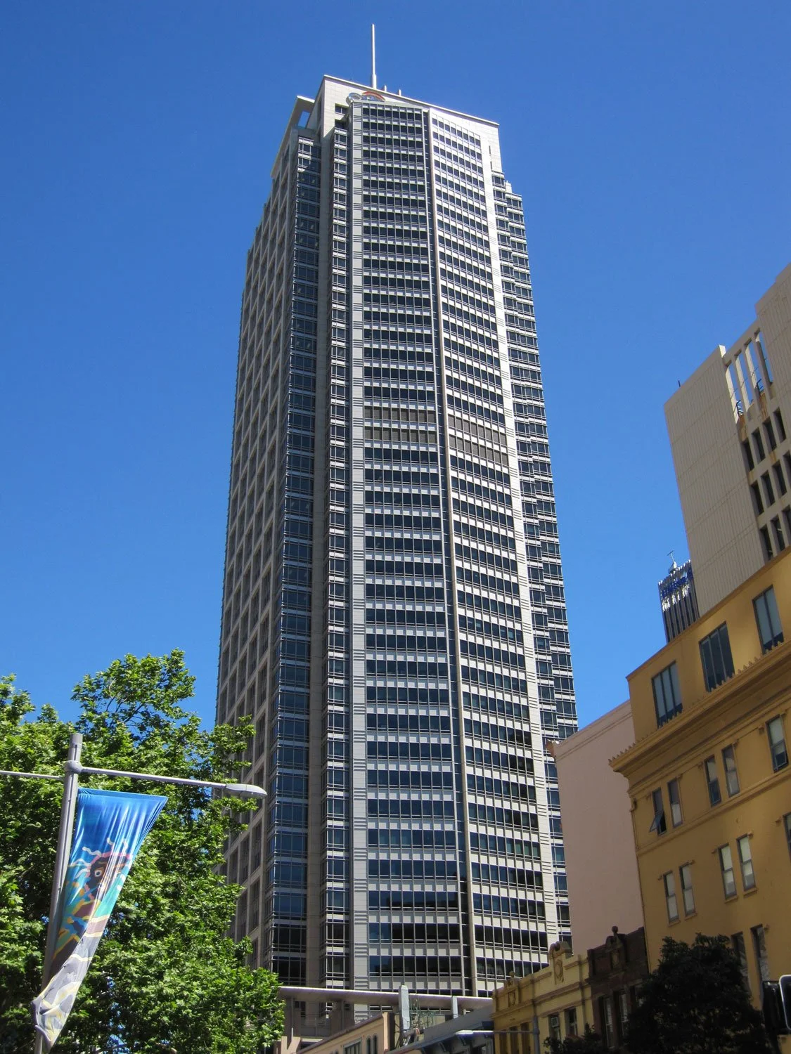 A tall modern skyscraper against a clear blue sky, surrounded by other buildings and greenery.