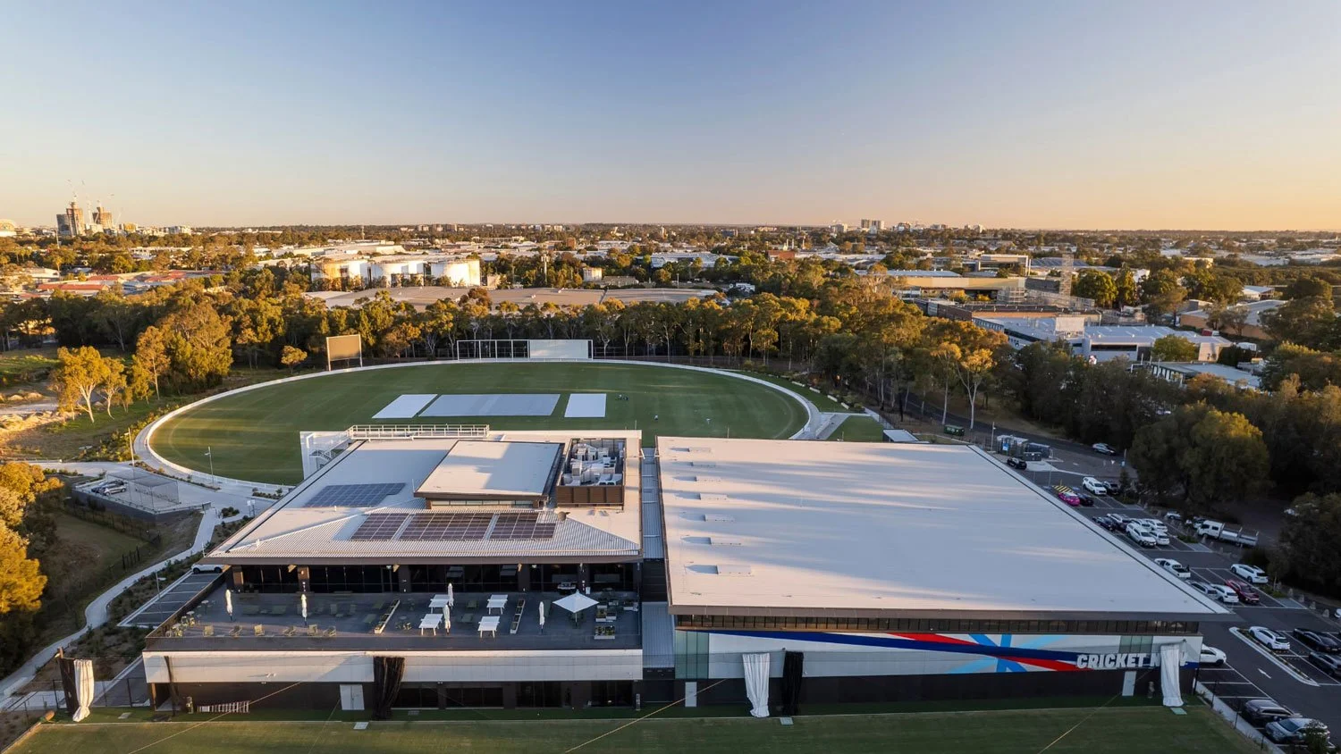 Aerial view of a sports complex with a cricket stadium, a running track, and adjacent buildings, surrounded by trees and parking lot, under a clear sky during sunset.