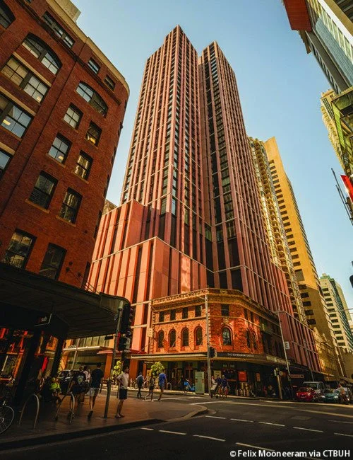 Tall pink and red skyscraper in a city with surrounding buildings, people walking on the street, and a clear blue sky.