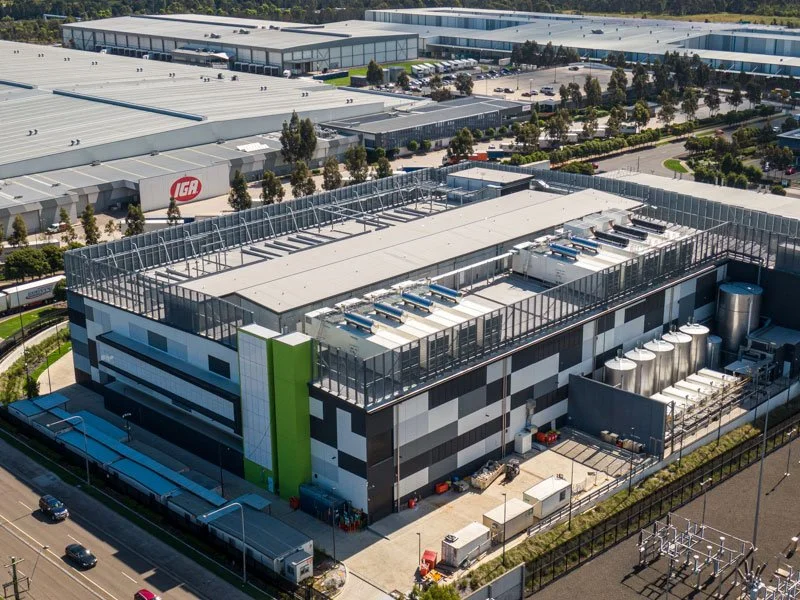 Aerial view of a modern industrial building with multiple stainless steel tanks and HVAC units on the roof, surrounded by parking lots and other similar warehouses.