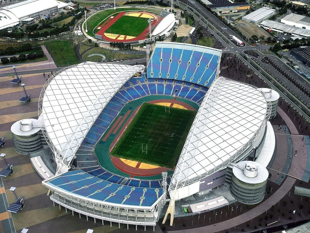 An aerial view of a large stadium with a green field, surrounded by seating and partial roofing, located near a running track, with nearby roads, parking lots, and a sports complex in the background.