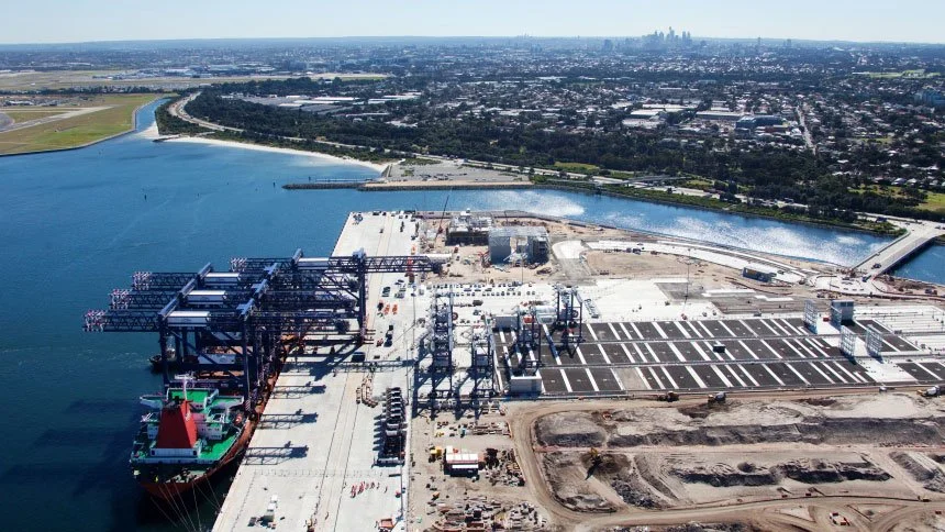 Aerial view of a large industrial port with a container ship docked, storage tanks, and construction areas, near a body of water and a city skyline in the distance.