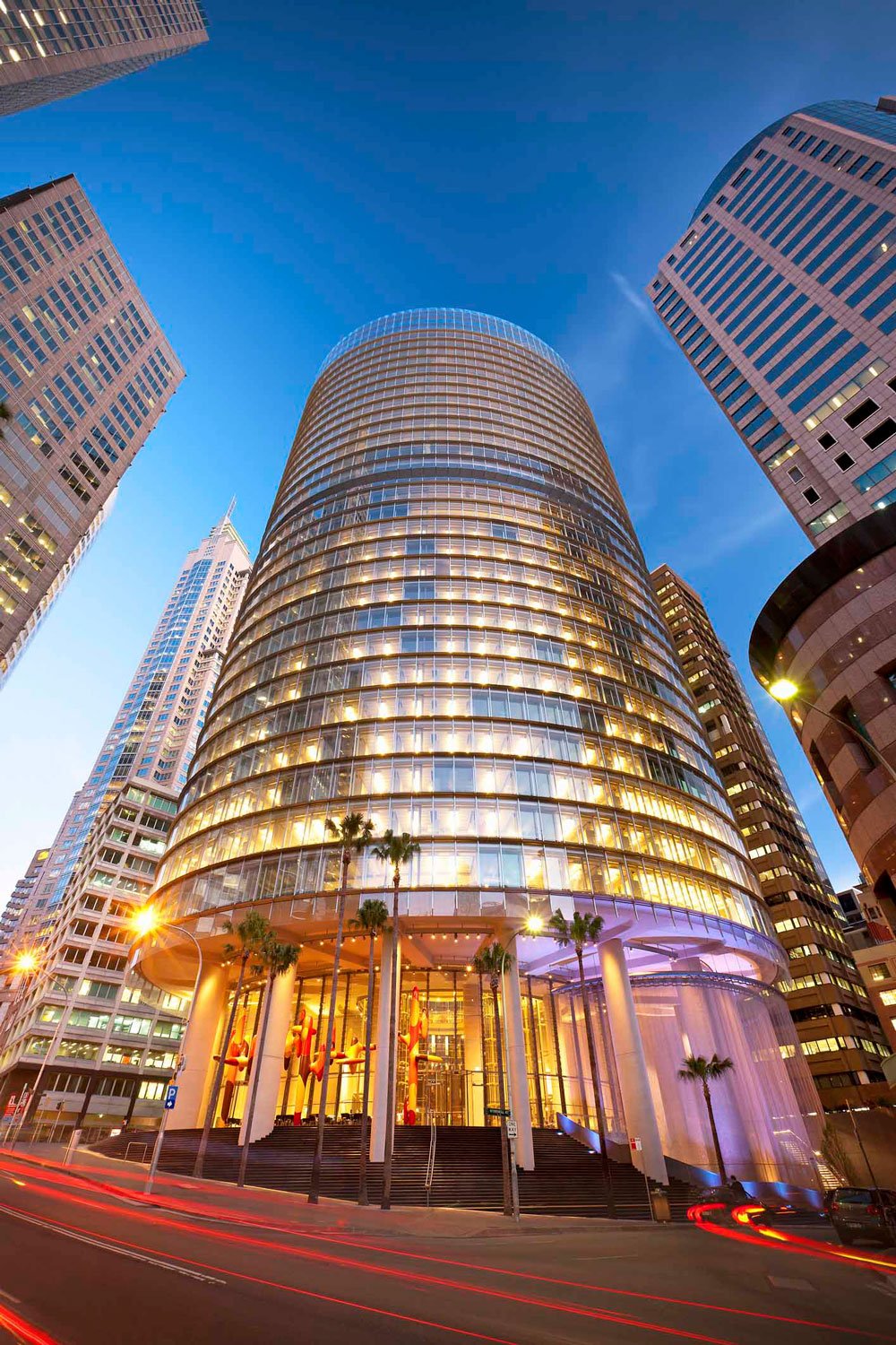 A tall modern skyscraper with a rounded glass facade, illuminated from within with warm yellow lights, surrounded by other high-rise buildings in a cityscape at dusk. Palm trees are at the base, and light trails from moving vehicles are visible on the street.