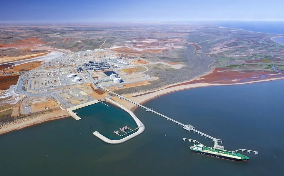 Aerial view of a nuclear power plant near a body of water with a dock, connected to a large cargo ship.
