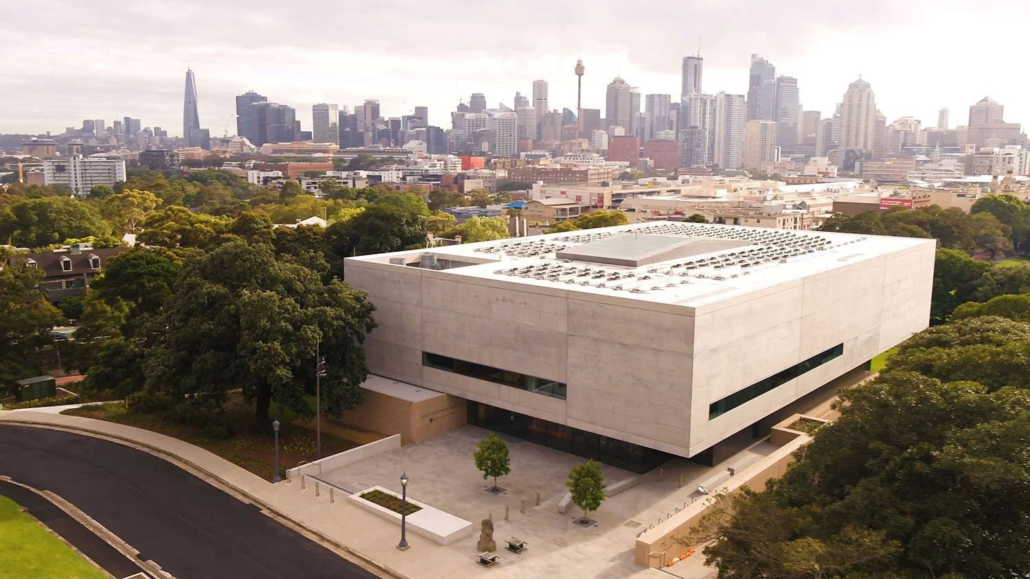 An aerial view of a modern white building surrounded by trees, with a city skyline featuring tall skyscrapers in the background.