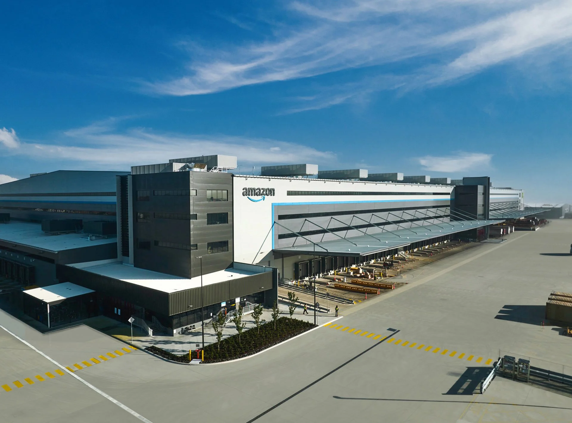 Exterior view of an Amazon distribution center with loading docks, a large parking area, and clear skies.