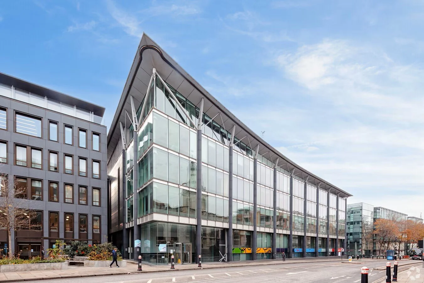 Modern glass building with sloped roof on a city street, with trees and parked cars.