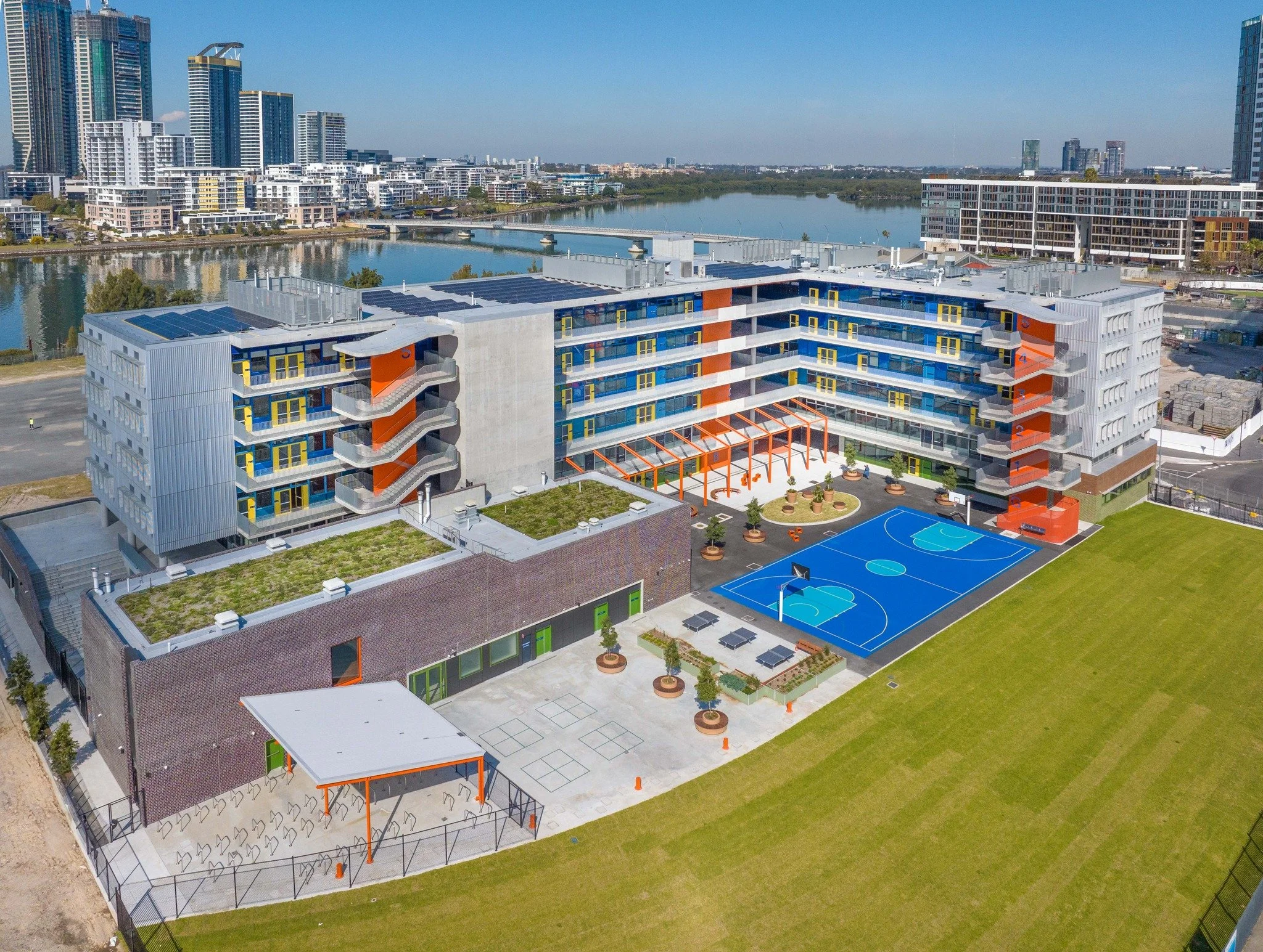 A modern school building with multiple floors, outdoor basketball courts, and a green sports field. The building has a colorful, contemporary design with orange accents. In the background, a river and city skyline are visible.