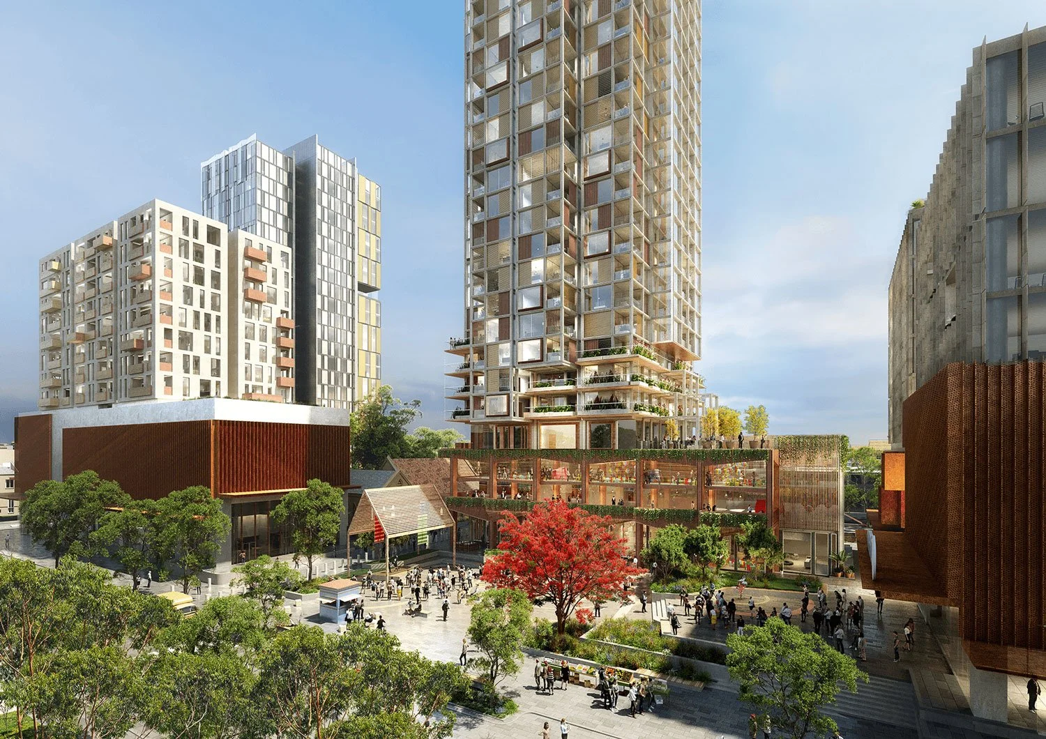 A modern urban development with tall residential buildings, a landscaped public plaza with trees, seating areas, and a crowd of people walking and gathering. A distinctive red tree adds color to the scene.