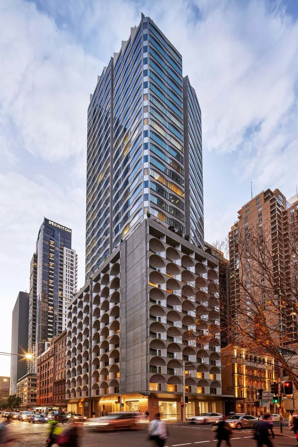 Tall modern skyscraper with unique curved balconies at the bottom and glass windows at the top, in an urban city with traffic and other tall buildings.