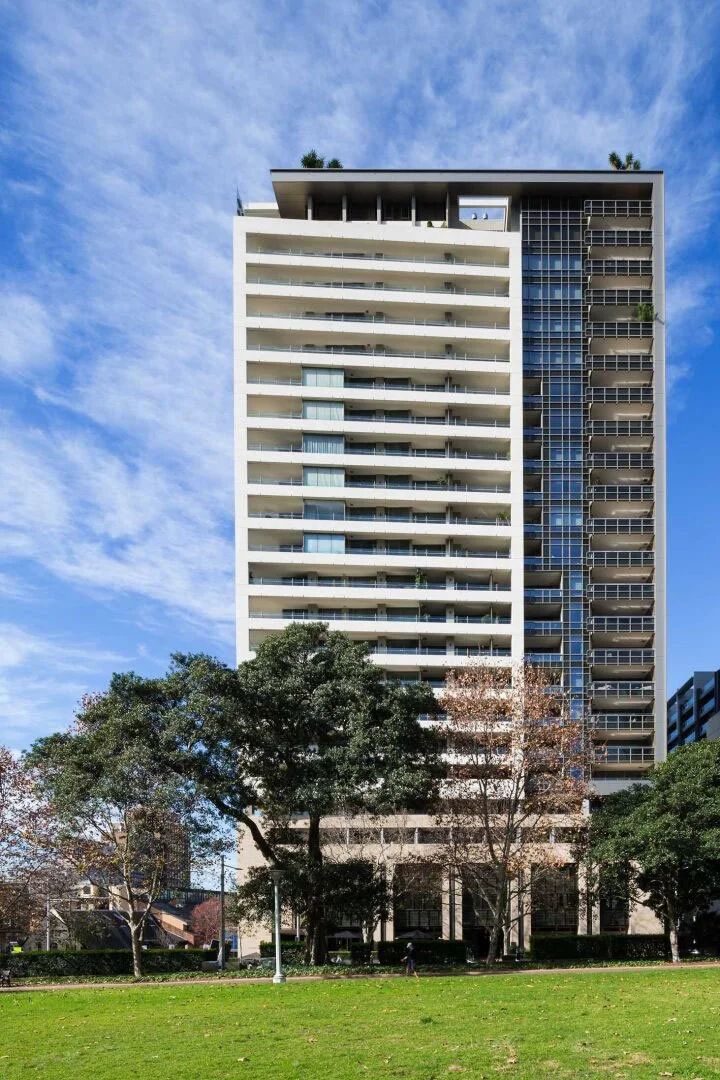 A tall modern residential building with multiple balconies and large glass windows, surrounded by trees and greenery, against a partly cloudy sky.