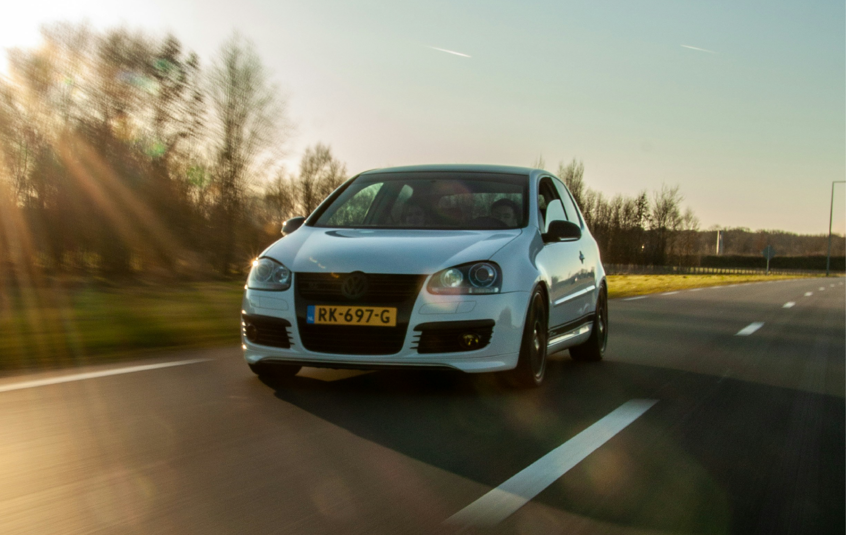 A white Volkswagen Golf driving on a highway during sunset with a blurred background of trees and sky.