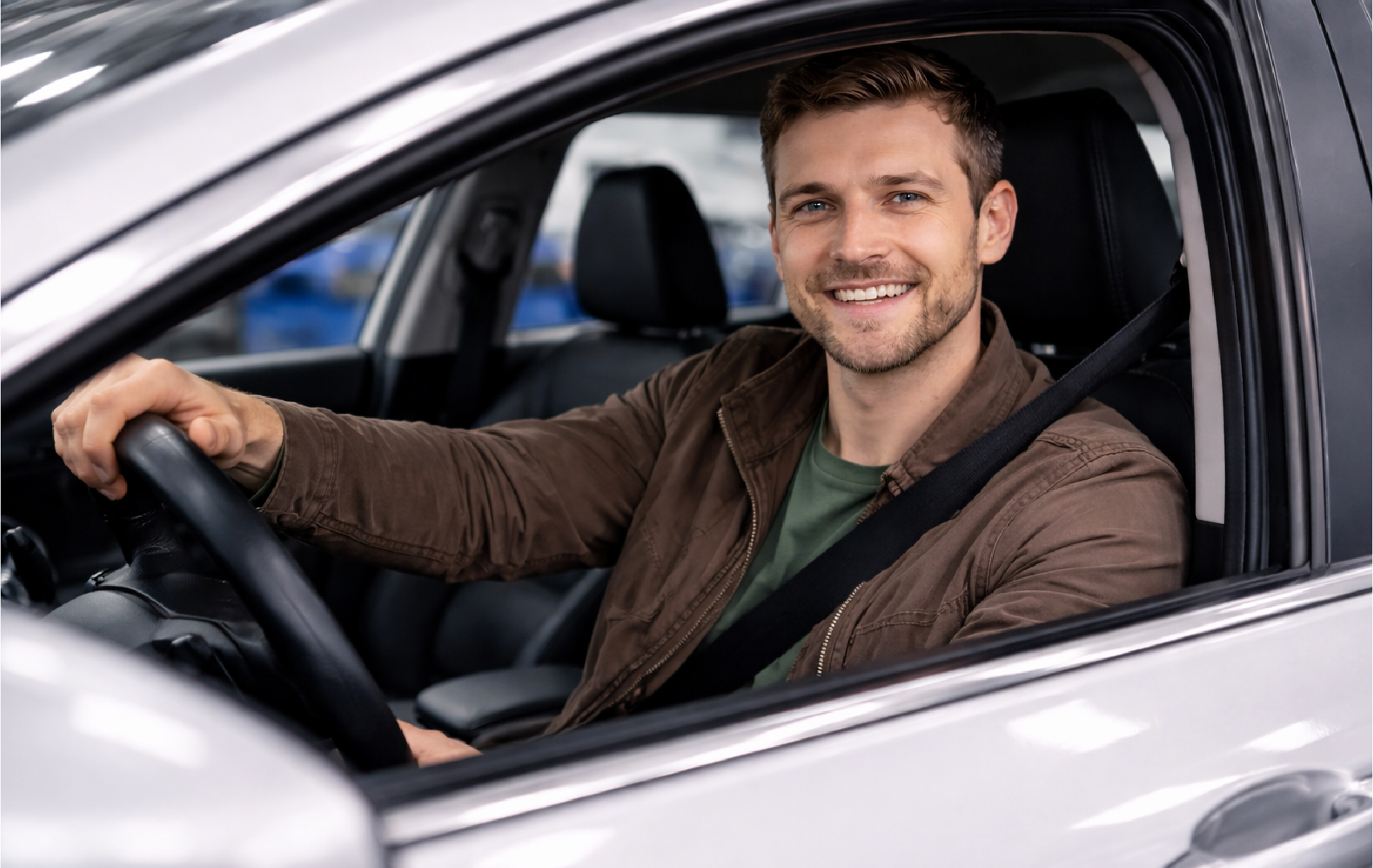 A smiling man with short brown hair and a beard driving a silver car, wearing a brown jacket and a seatbelt inside a parking garage.