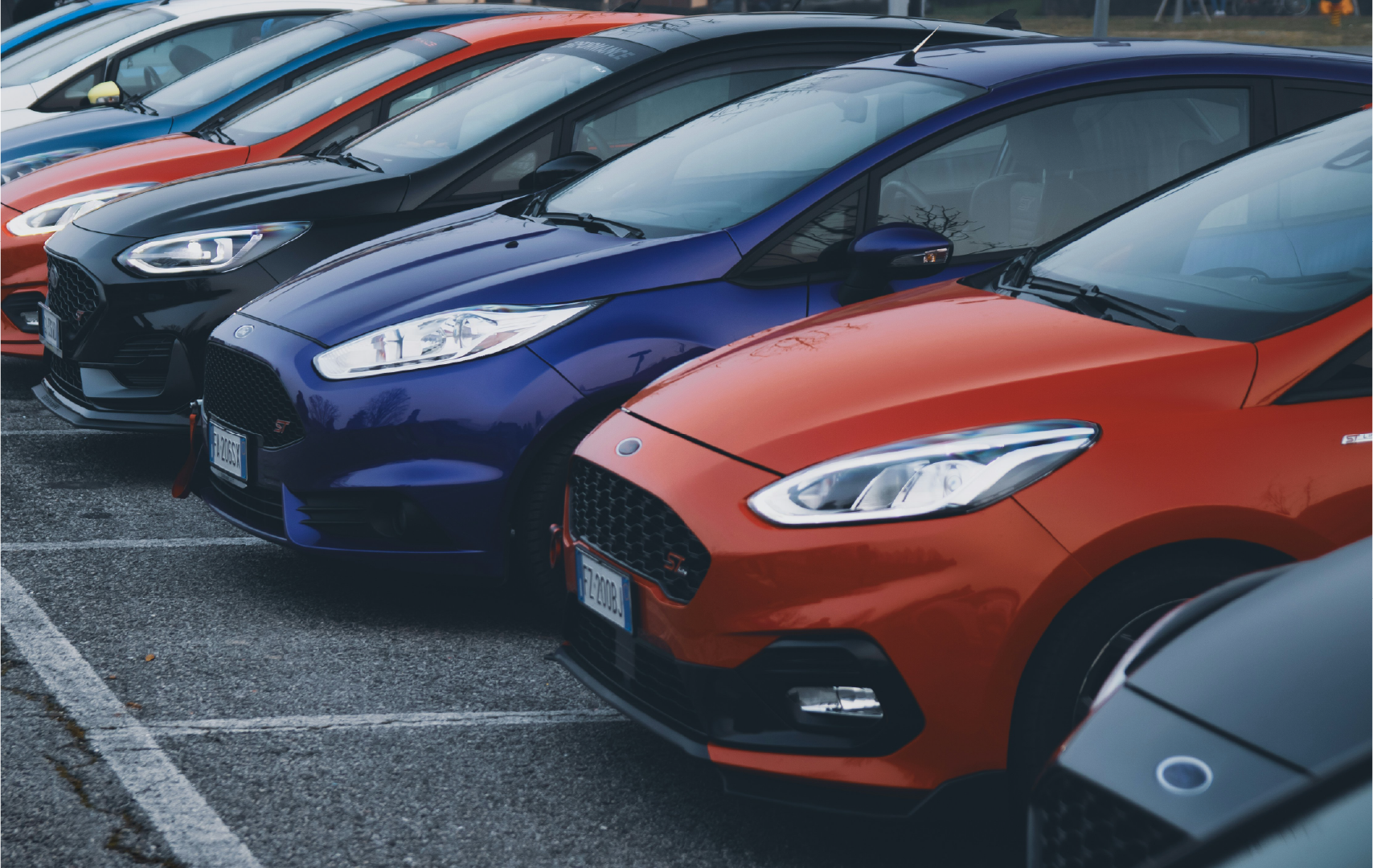 Several sporty cars parked in a parking lot, with a black, blue, orange, and gray car at the front.