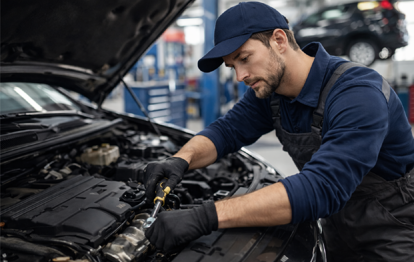 A male mechanic wearing a navy uniform and cap working under the open hood of a car in a garage or auto repair shop.