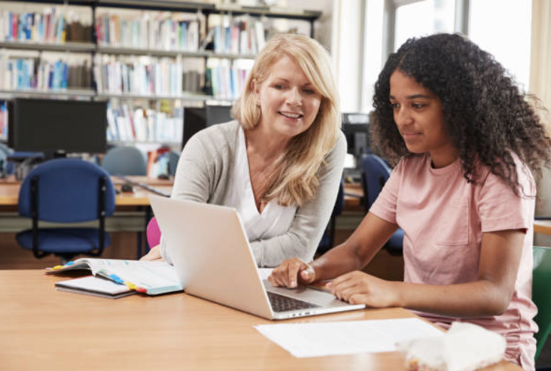 A teacher and student working together at a desk in a library, with bookshelves and computers in the background.