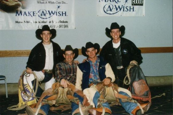 Four young men dressed in cowboy attire, including hats and boots, posing together in front of Make-A-Wish banners at an indoor event.