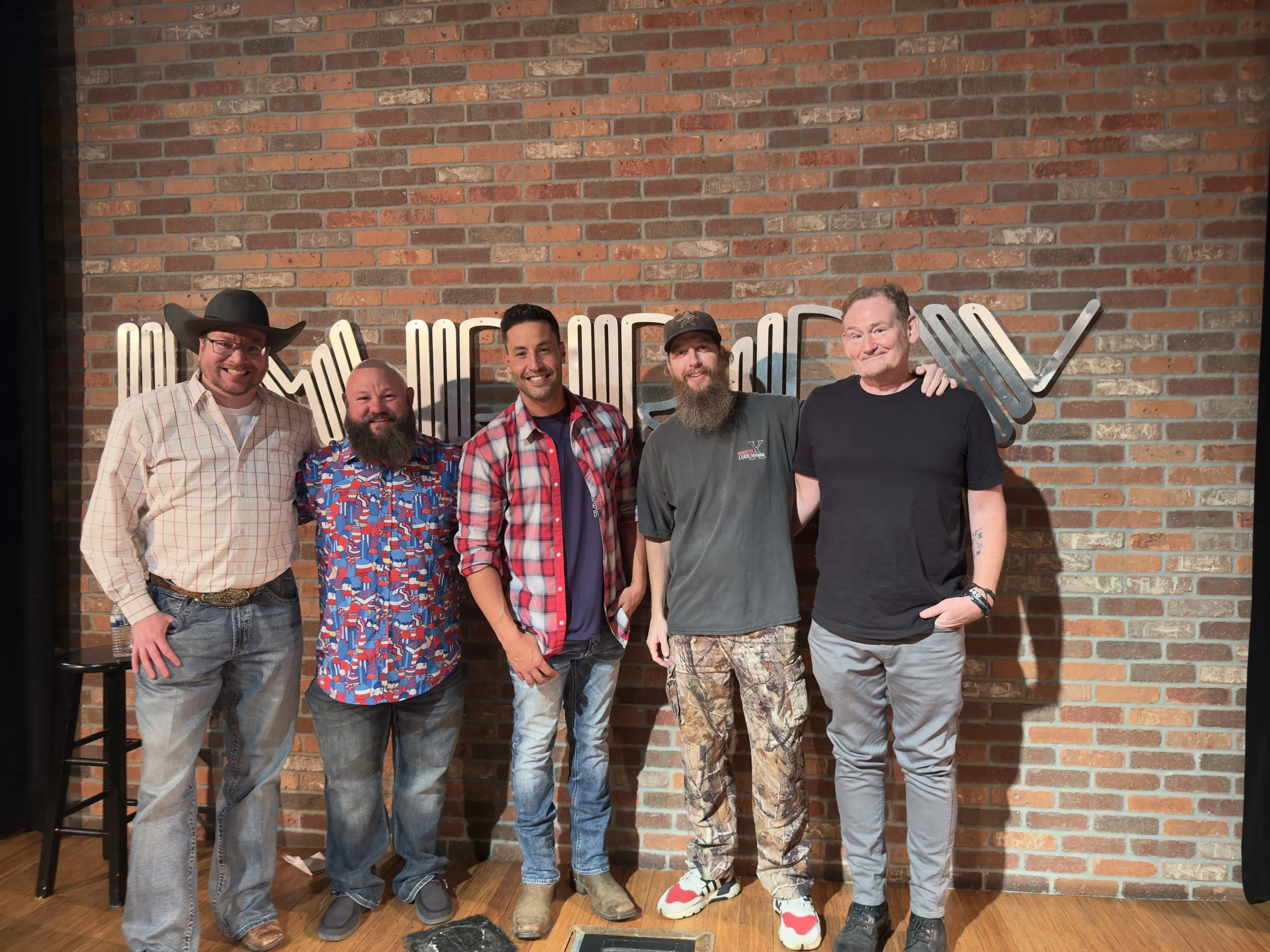 Five men standing together in front of a brick wall, smiling. One man wears a cowboy hat, another has a colorful patterned shirt, the third wears a plaid shirt, the fourth has a beard and wears a gray t-shirt and cap, and the fifth wears a black t-shirt and gray pants.