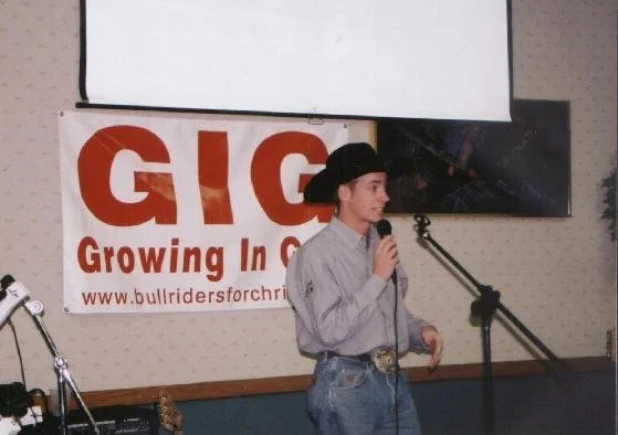 A woman wearing a cowboy hat and gray shirt speaking into a microphone at an indoor event with a GIC banner in the background.