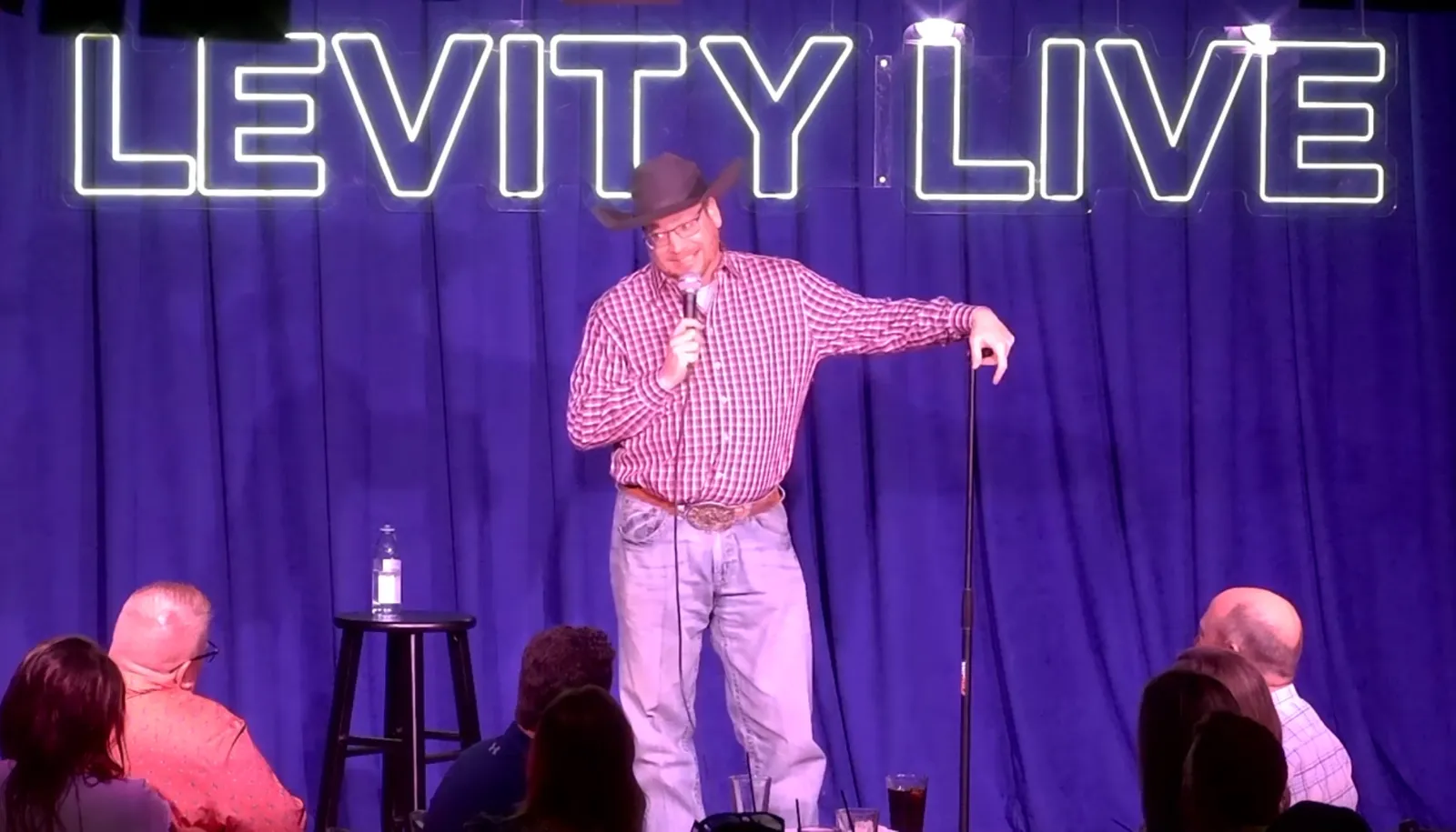 A comedian on stage wearing a cowboy hat, plaid shirt, and jeans, holding a microphone, with a neon sign behind him that says 'LEVIY LIVE' and an audience sitting in front of him.