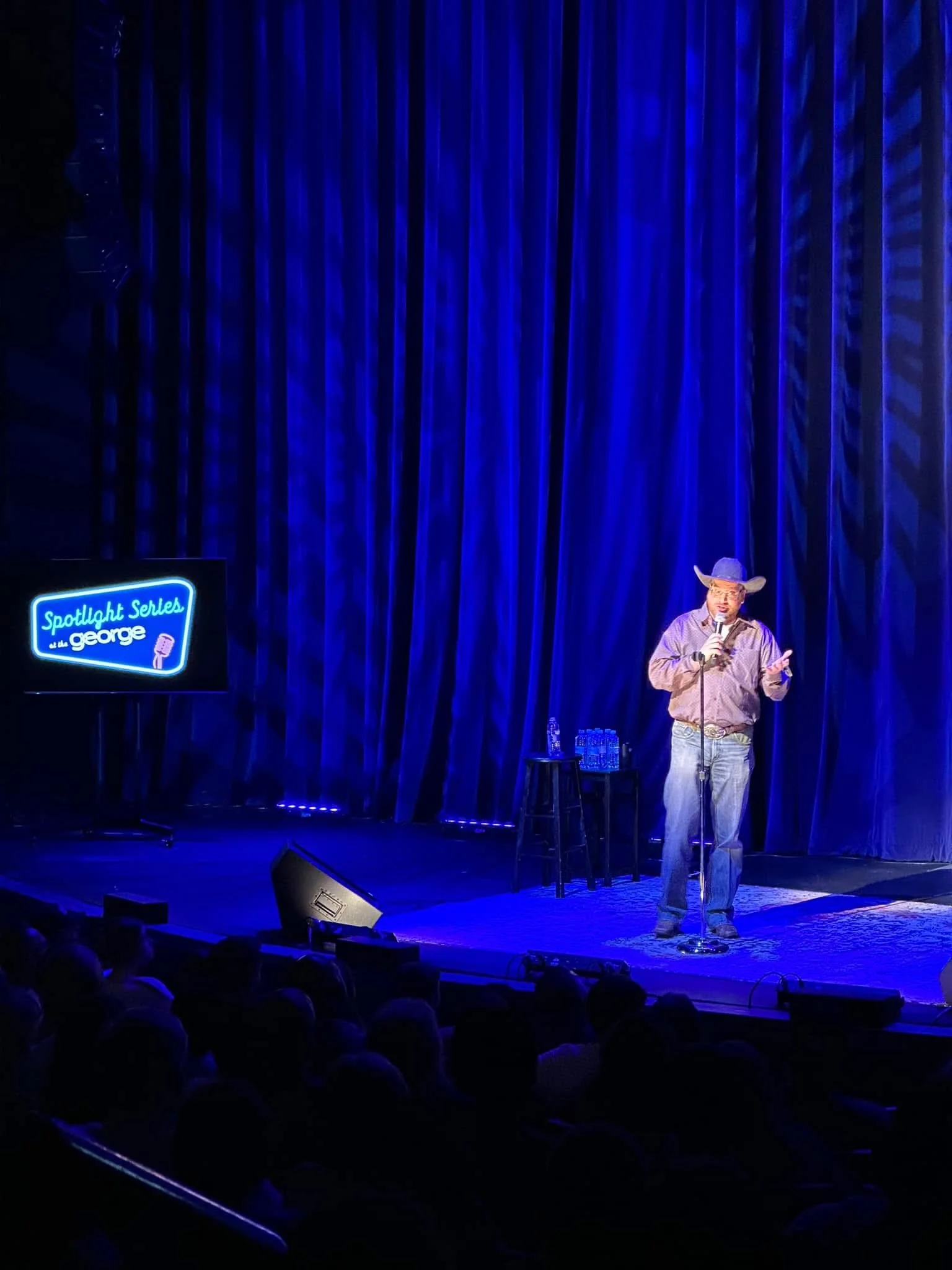 A comedian wearing a cowboy hat performing stand-up on stage with blue curtains in the background at the 'Spotlight Series' in George, with a small table holding water bottles and a stool nearby.