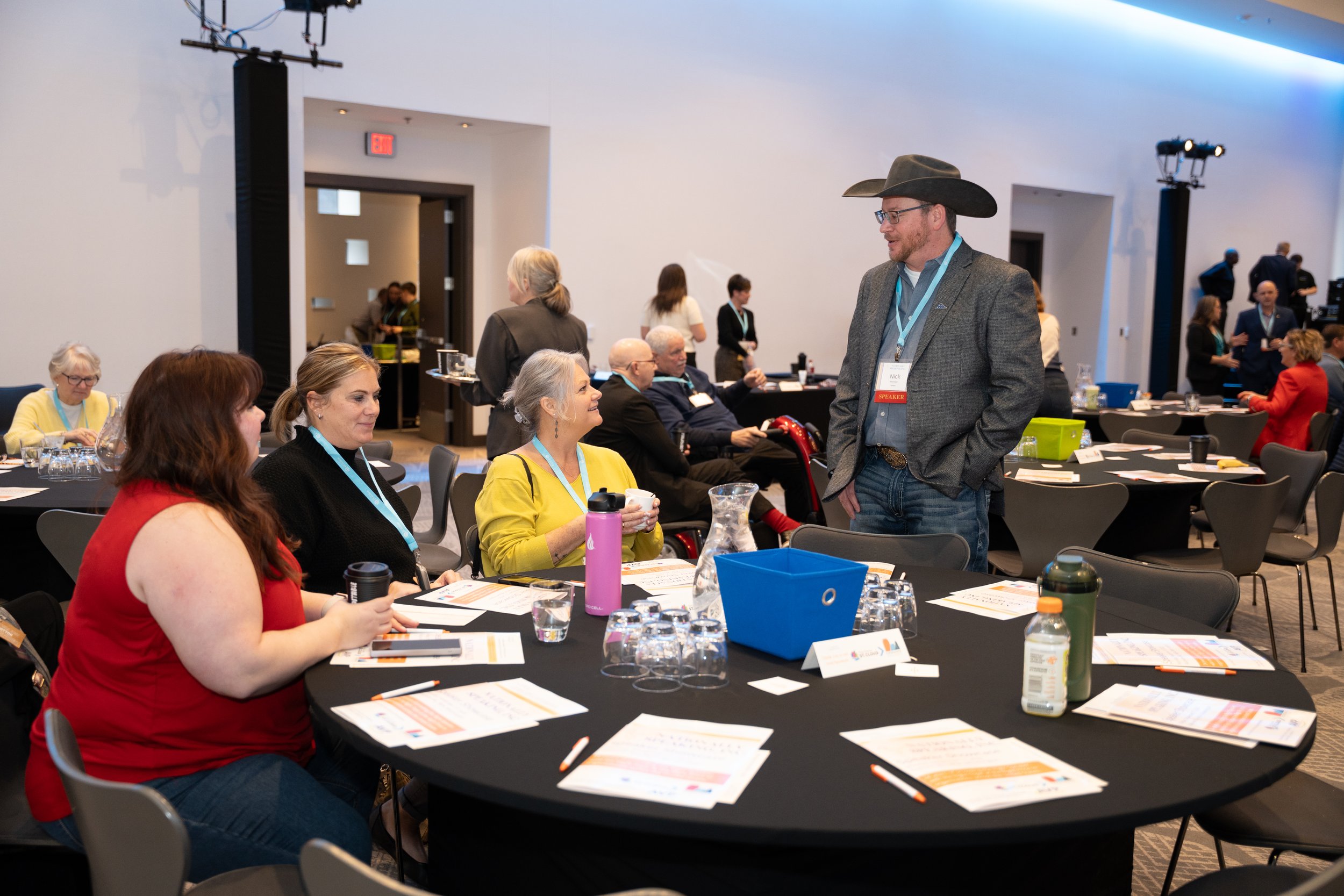 A man wearing a cowboy hat and gray blazer is talking to three women sitting at a round conference table. The table has papers, pens, water glasses, and reusable bottles. Other attendees are visible in the background in a conference setting.