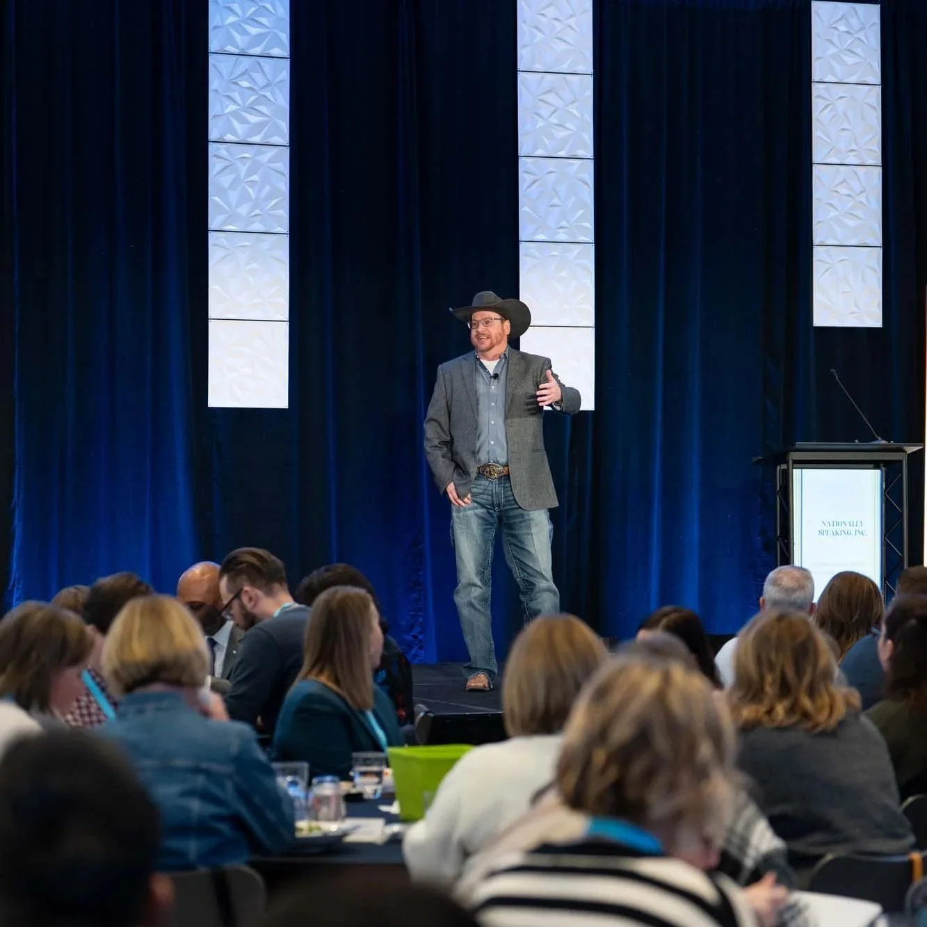 A man wearing a cowboy hat and blazer giving a presentation on stage at a conference, with an audience seated in front of him.