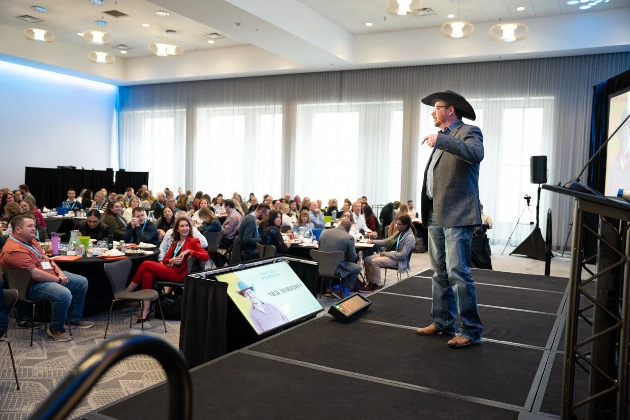A man wearing a cowboy hat and blazer giving a presentation to a large audience in a conference room with high ceilings and large windows.