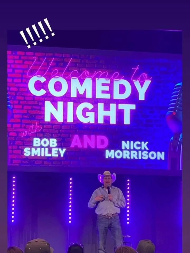 A man wearing a cowboy hat and jeans speaking into a microphone on stage at a comedy night event, with a large neon sign behind him that reads 'Welcome to COMEDY NIGHT with Bob Smiley and Nick Morrison' in bright neon colors. Audience members are visible in the foreground.