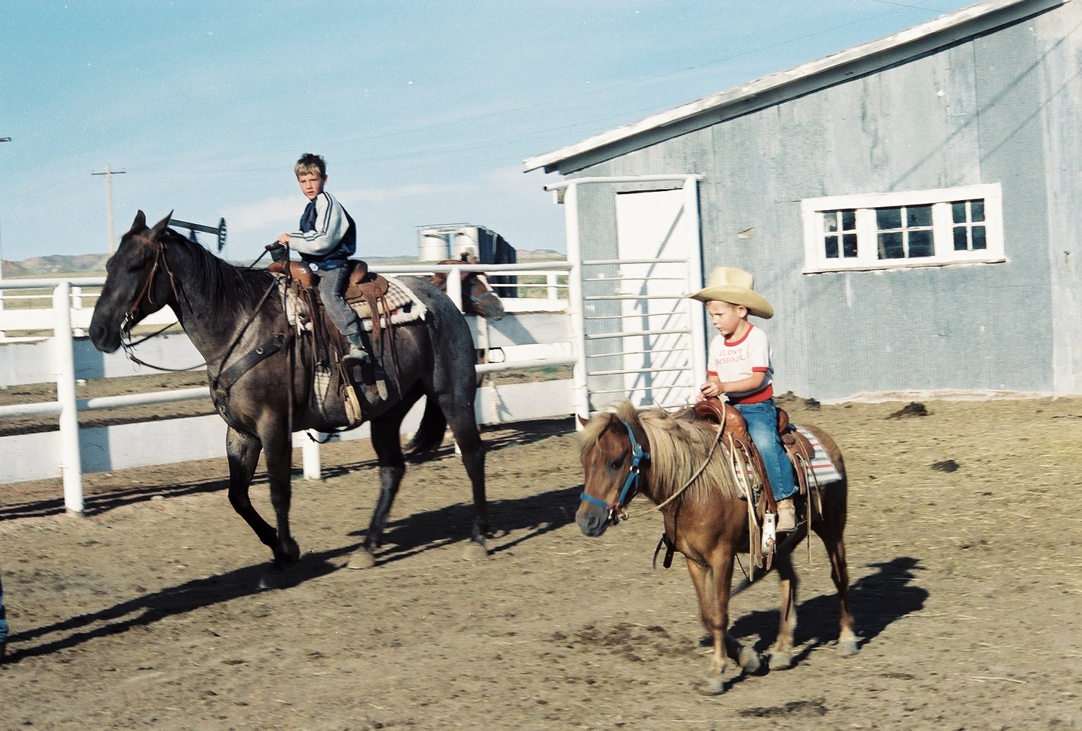Two young children riding ponies in a dirt paddock. The boy is on a larger dark horse, and the girl on a smaller light brown pony, both wearing cowboy hats and casual clothing.