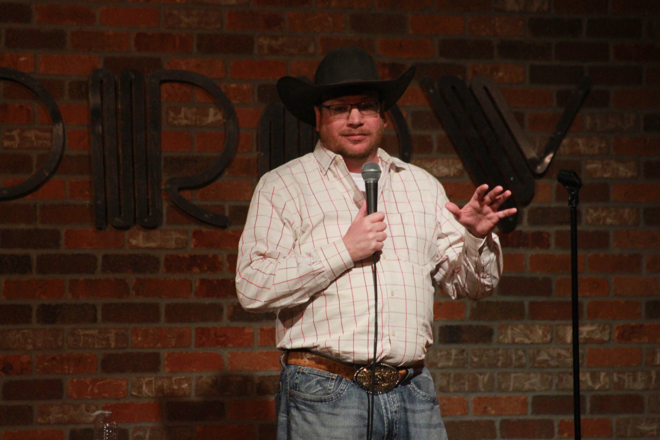A man dressed as a cowboy stands on stage in front of a brick wall with the word 'Comedy' spelled out in large black letters. He is wearing a black cowboy hat, glasses, a plaid shirt, jeans, and a belt with an ornate buckle, holding a microphone, and appears to be performing stand-up comedy.